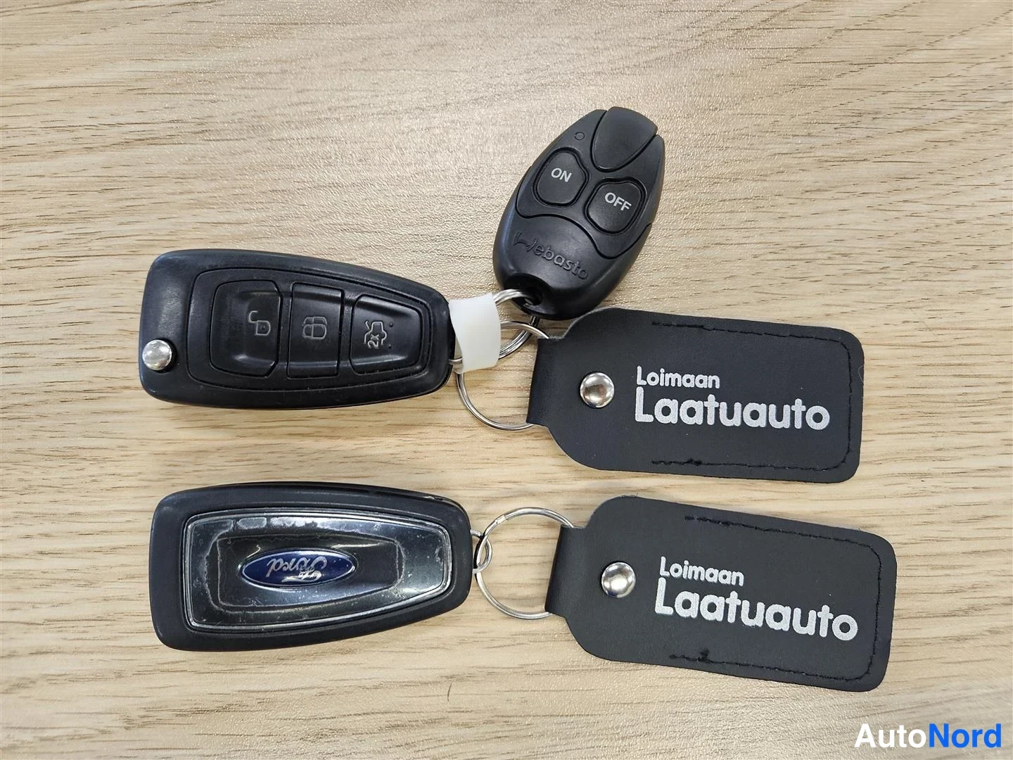 Yksityiskohta: Kuva auton avaimista: kaksi avainpainiketta, lohkolämmitin-/alarmikauko-ohjain ja kaksi Loimaan Laatuauto -avainnappia puupintaisella pöydällä.