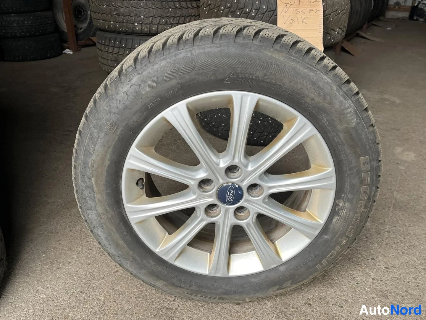 Yksityiskohta: Hopeinen levyjalka Ford-merkkipainikkeella ja nastarenkaisi kevytmetallivanteeseen kiinnitettynä, taustalla renkaita ja korte.