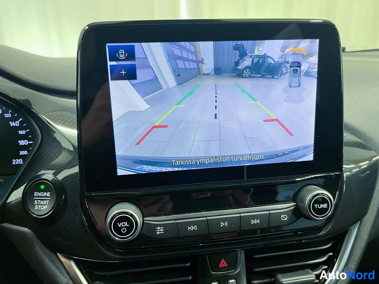 Muu: Infotainment-näyttö näyttää peruutuskameran kuvaa huoltamo-hallista ohjausviivoineen ja tekstillä 'Tarkista ympäristön turvallisuus'.