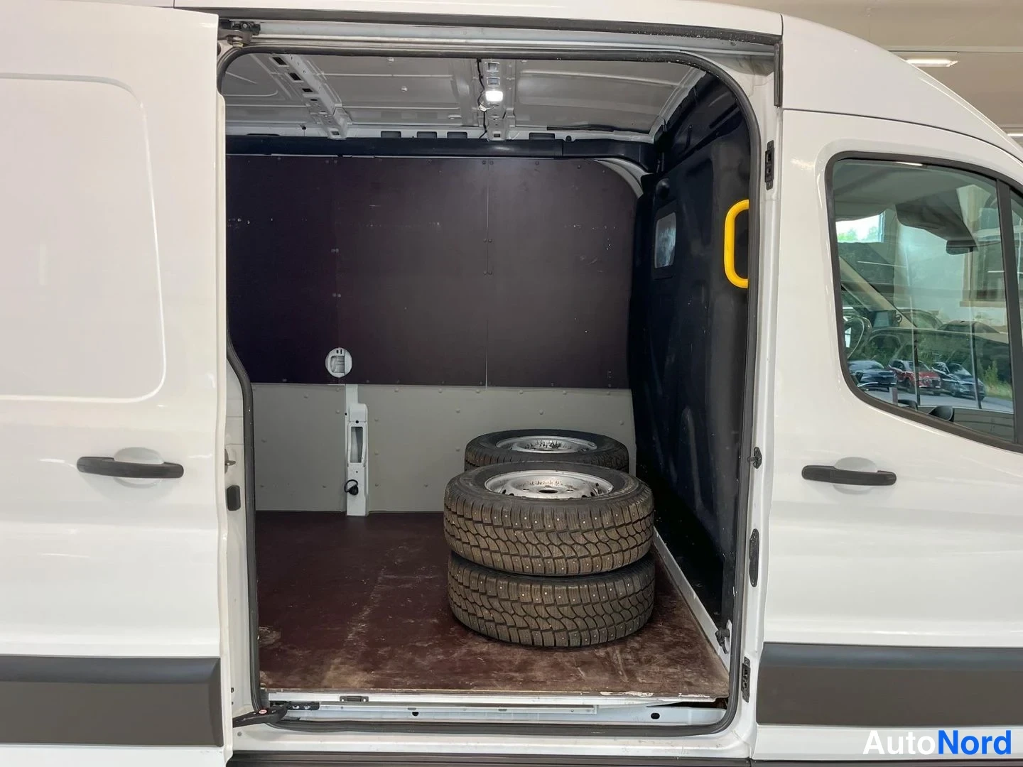 Takaa: Ford Transit -pakettiauton takaosan lastitila, takaovet auki. Lattialla on kolme pinoa nostettua nastarenkaita vanteineen. Sisäpanelit tummaa ja vaaleaa, korin