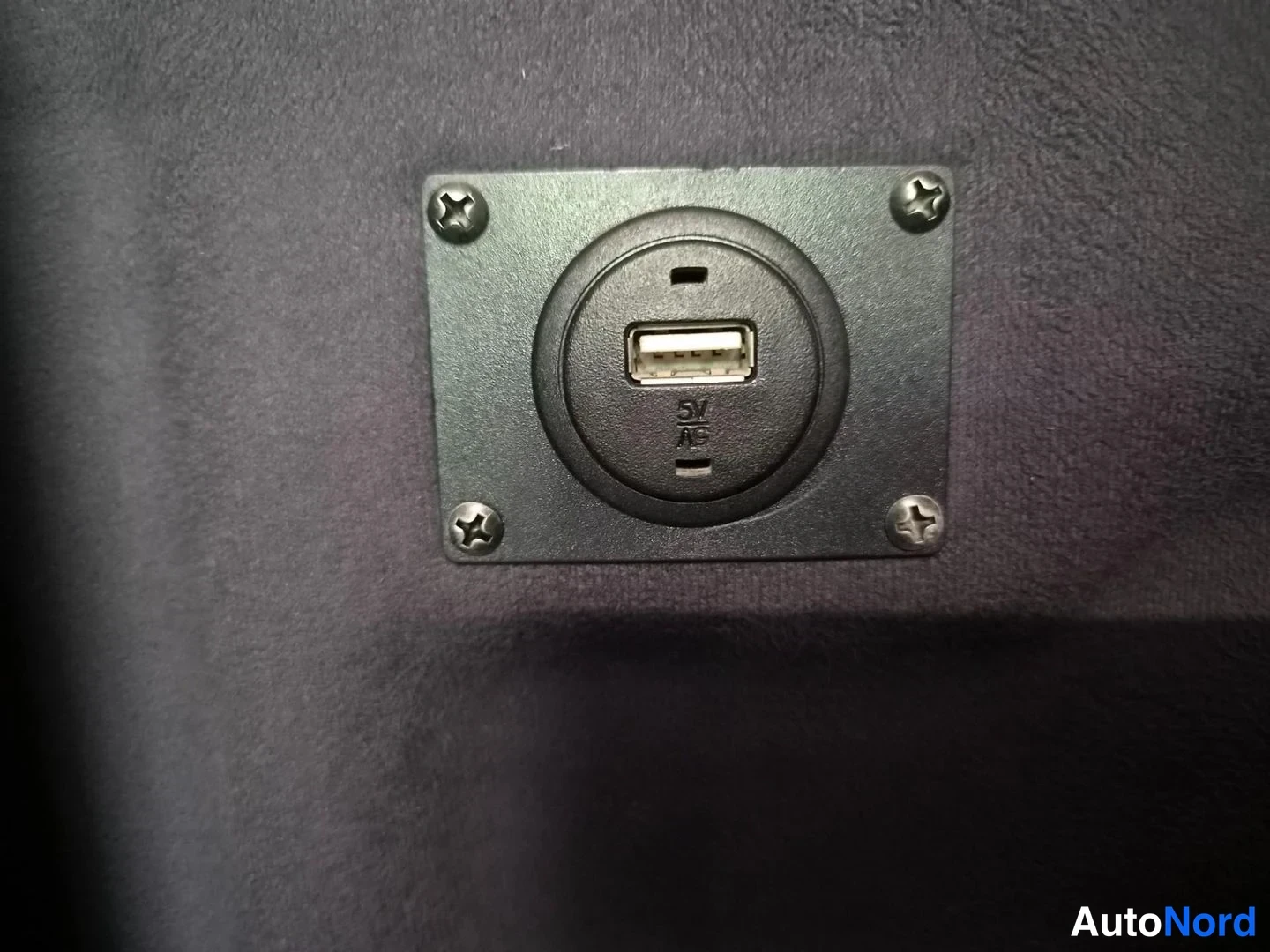 Yksityiskohta: Tummaan panelointiin asennettu pyöreä USB-portti, jonka ympärillä merkintä 5V ja 1A ja ruuvikiinnitys