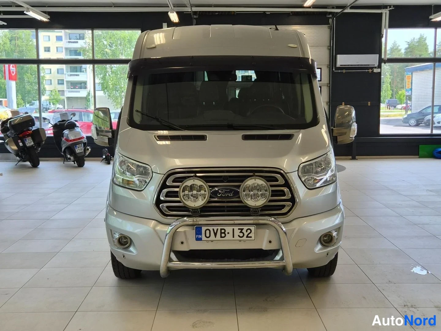 Edestä: Hopeanvärinen Ford Transit -pakettiauto suoraan edestä, etusäleikössä Ford-merkki ja kaksi lisävaloa, rekisteritunnus.