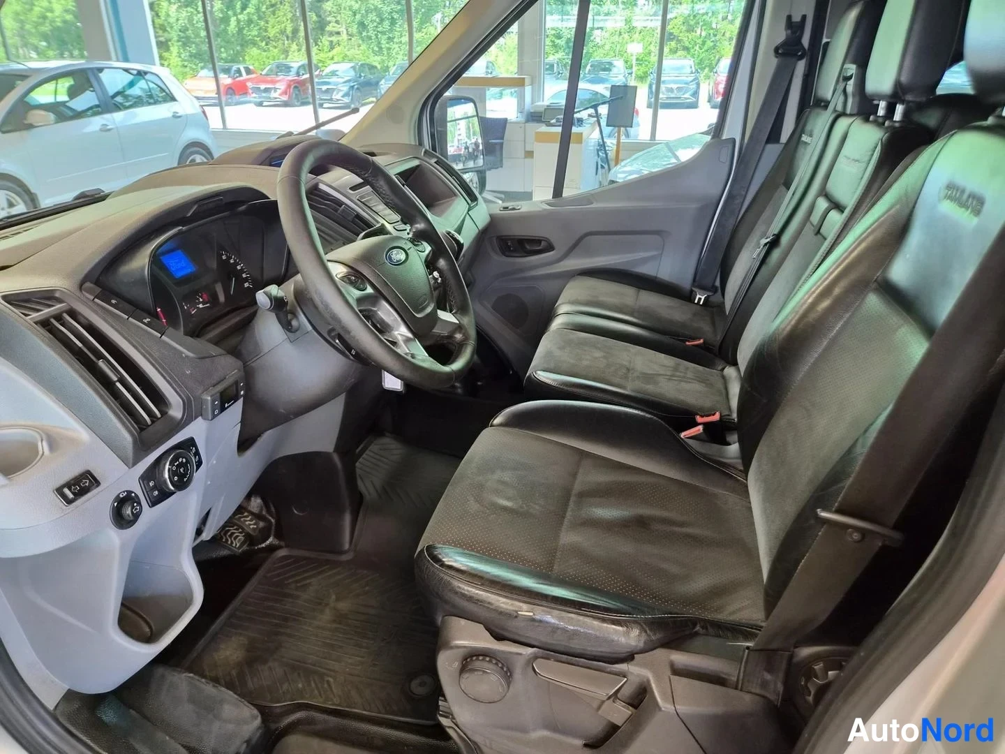Muu: Ford Transitin ohjaamo vasemmalta: ratissa Ford-merkki, mittaristo, tummat istuimet ja penkkirivin turvavyöt