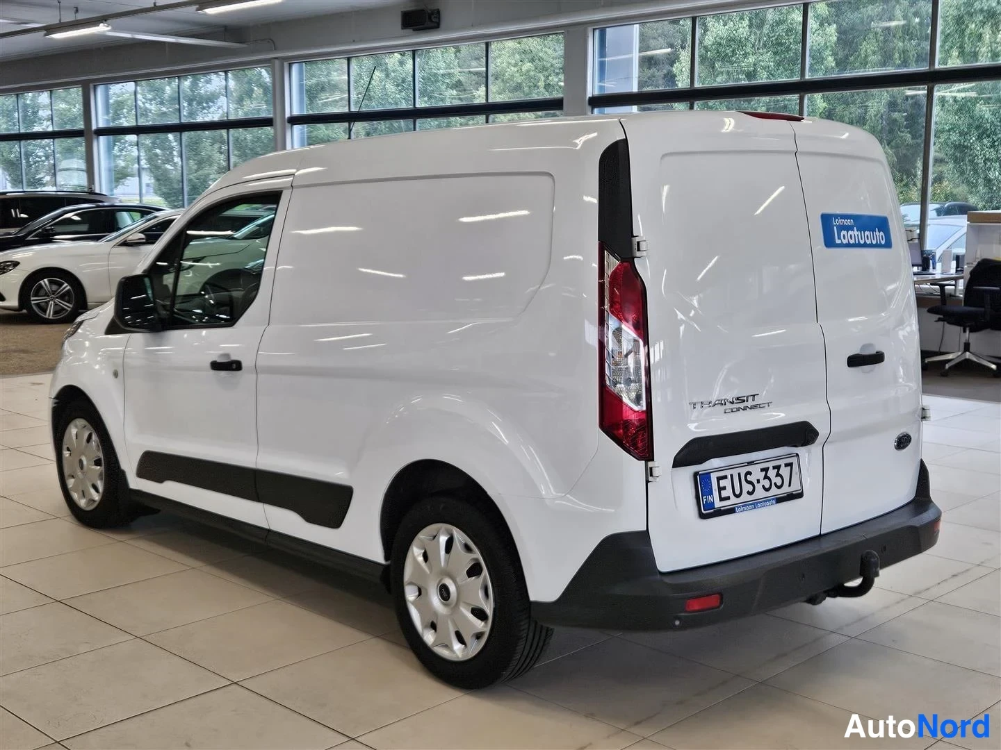 Takaviisto: Ford Transit Connect -pakettiauton takakolmasosa ja sivu näkyvissä valkoisena lasiseinäisessä autosalissa; kilpi ja Transit Connect -merkintä takaluukussa.