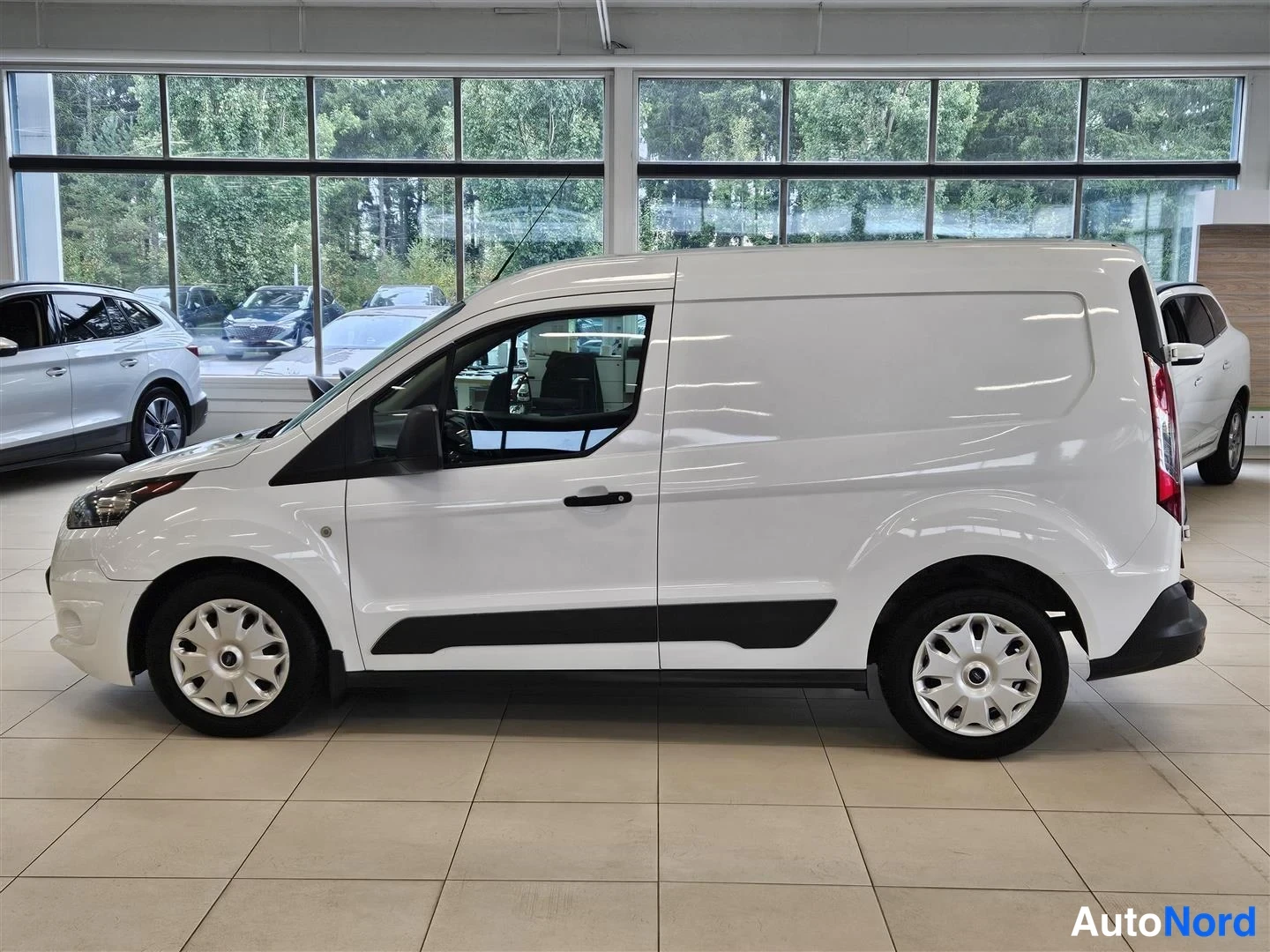 Etuviisto, nokka vasemmalle: Valkoisen Ford Transit Connect -pakettiauton sivukulma etupää kuvan vasemmassa laidassa, autosalin lattiassa ikkunaseinän edessä.