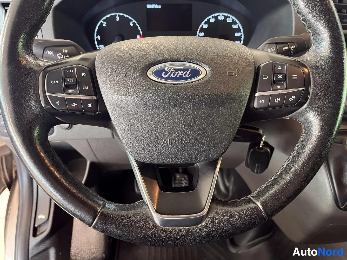 Yksityiskohta: Ohjauspyörän close-up: Ford-merkillinen ilmatyyny keskellä, nappiryhmät molemmin puolin (CAN, RES, SET-, LIM, OK) ja mittarit taustalla.