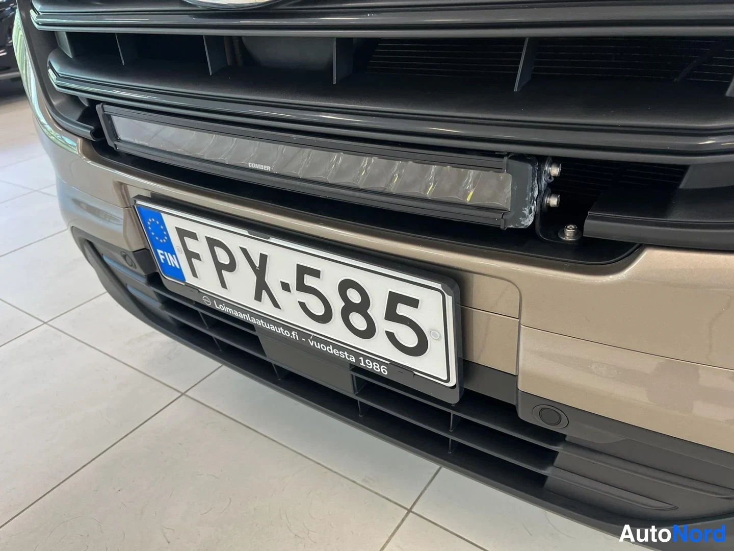 Edestä: Lähikuva Ford Transit Customin etupuskurista ja etusäleiköstä, jossa rekisterikilpi ja sen yläpuolella musta ajovalosarja.