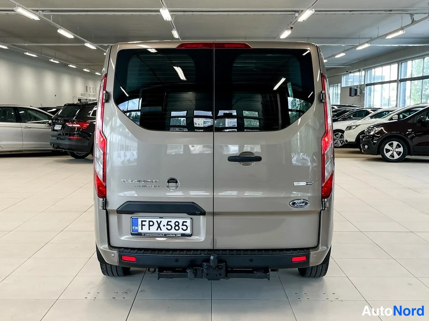 Takaa: Ford Transit Custom -pakettiauto takaa nähtynä autosalissa, beigen runko, rekisterikilpi ja takavalojen väliin asennettu Ford-tunnus.