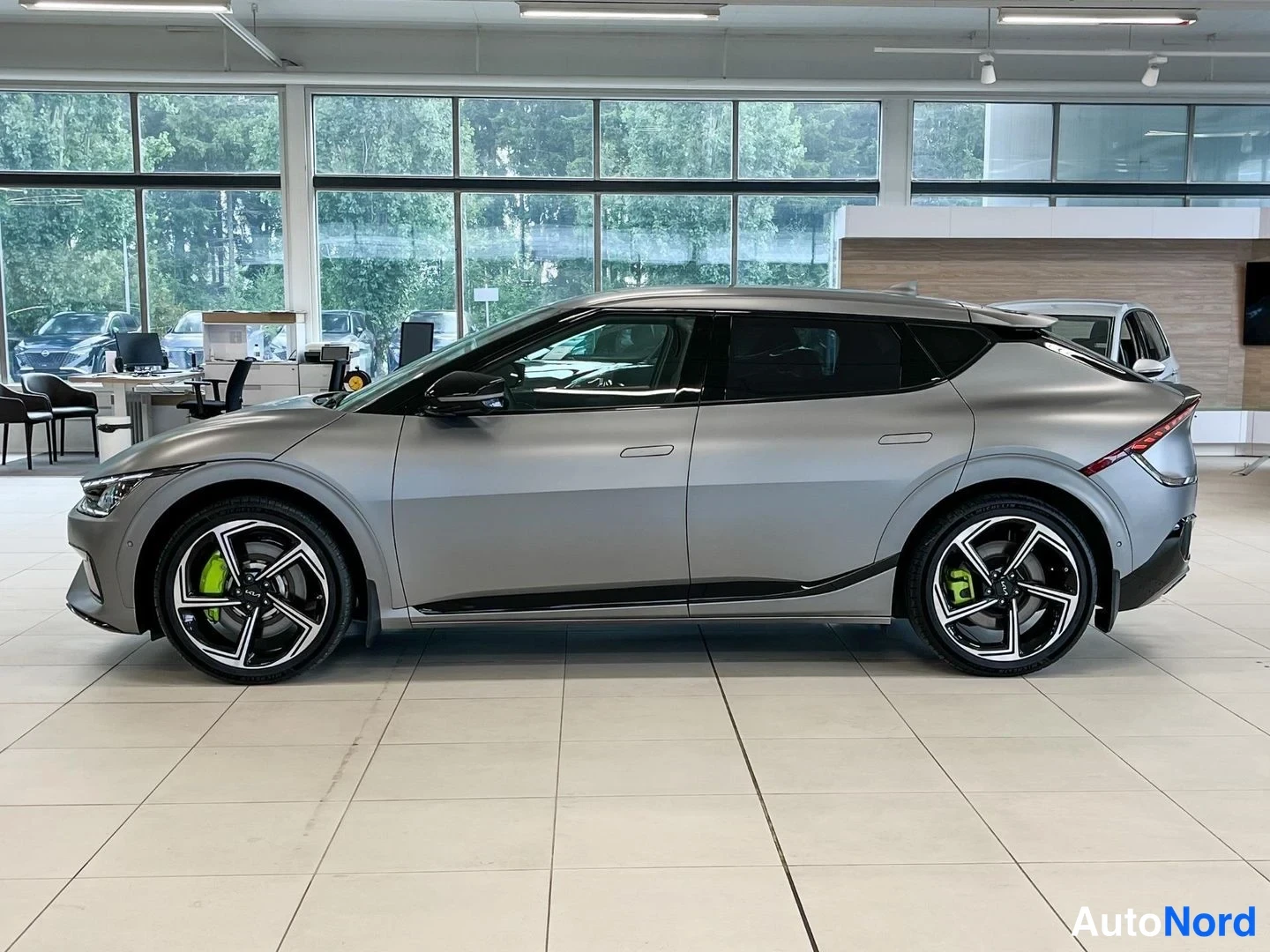 Etuviisto, nokka vasemmalle: Kia EV6:n sivuprofiili autosalissa, harmaa kori, viisivalo­spiritäiset mustat kevytmetallivanteet ja vihreät jarrusatulat.