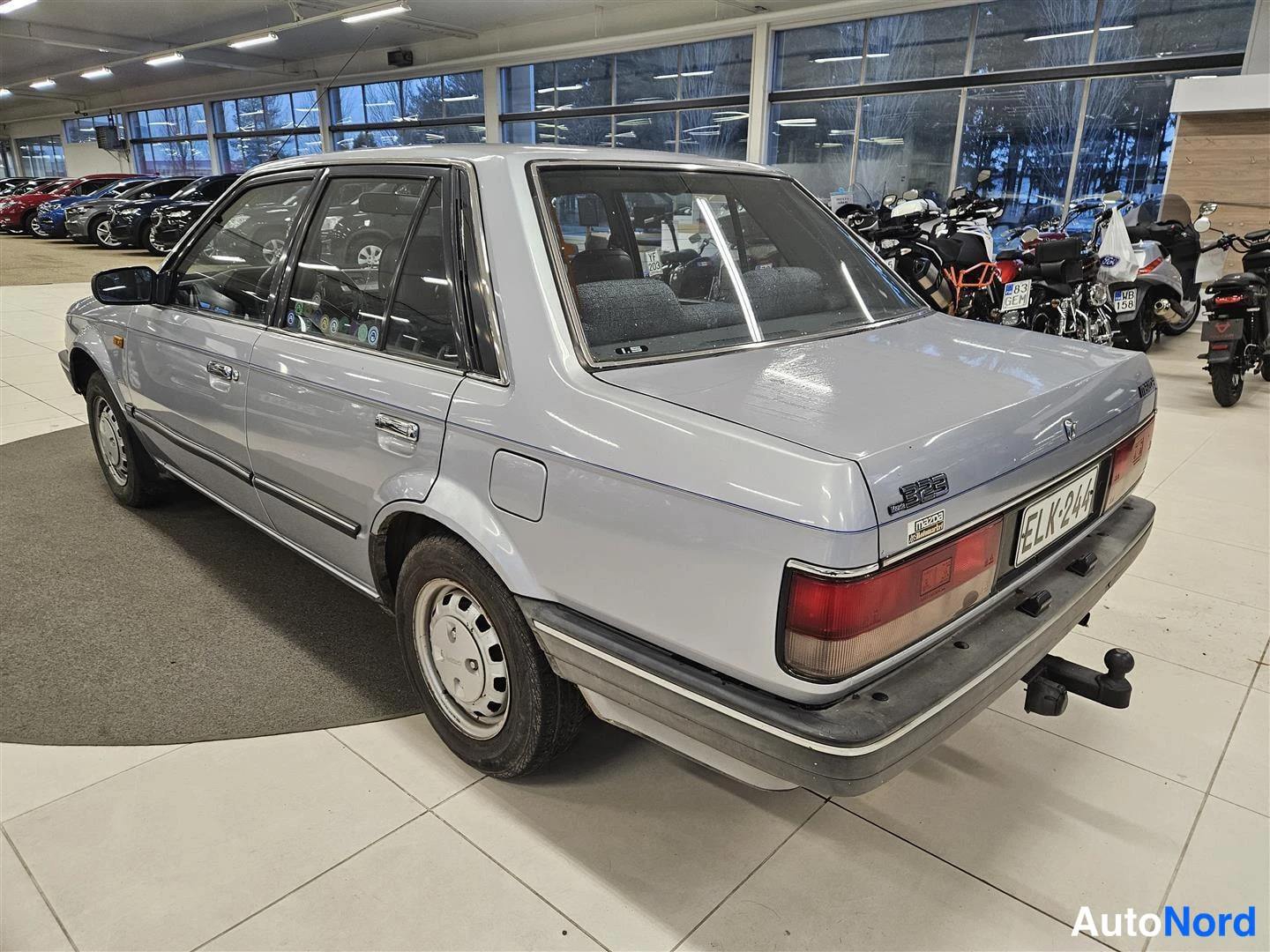 Takaviisto: Hopeanharmaa Mazda 323 -sedan takaa ja vasemmalla sivulta näkymässä, perävaunukoukku ja rekisterikilpi.