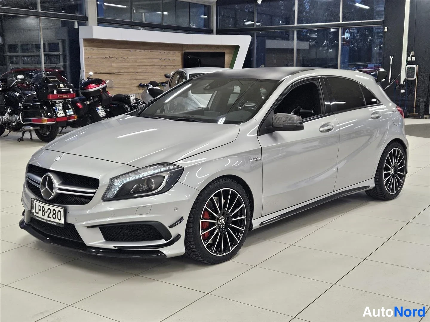 Etuviisto, nokka vasemmalle: Hopeanvärinen Mercedes-Benz A -mallinen henkilöauto kuvattuna etukulmasta autosalissa, mustat moniosaiset kevytmetallivanteet ja punaiset jarrusatulat