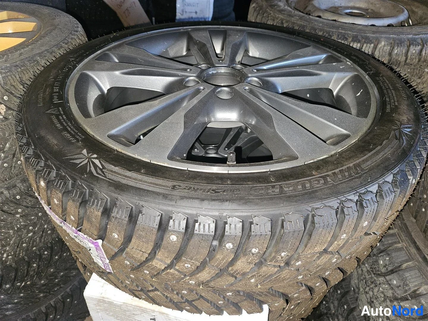 Muu: Musta pikkeli­rengas Nokian alumiinivanteella, renkaan sivussa lukee 'Nokian Tyres' ja 'WINGUARD', taustalla pino muita renkaita.