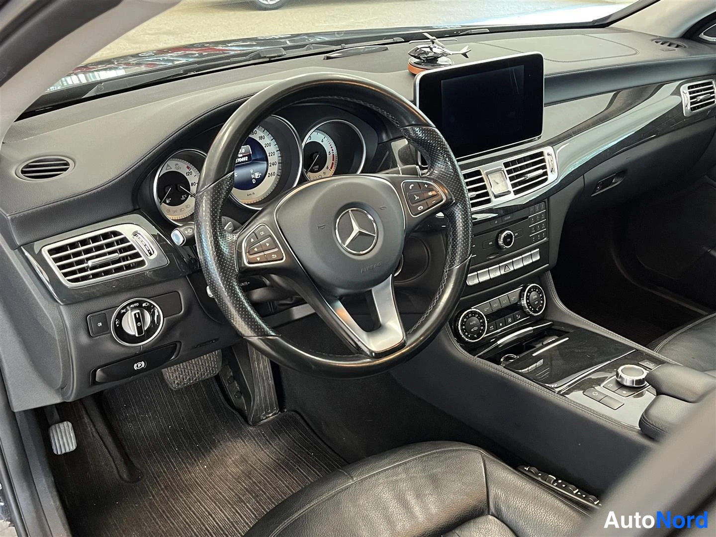 Edestä: Auton ohjaamossa näkyy Mercedes-Benz-ohjauspyörä, analogiset mittarit ja konsolin infotainment-näyttö.