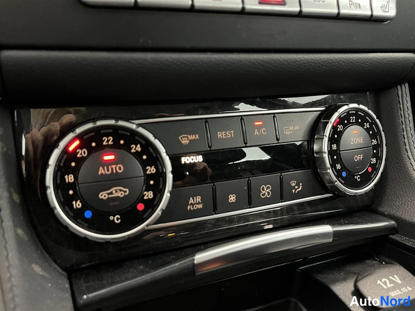 Yksityiskohta: Autohion ilmastointipaneeli kahdella pyöreällä säädöllä, näppäimet REST, A/C ja AIR FLOW sekä 12V-pistorasian kansi.
