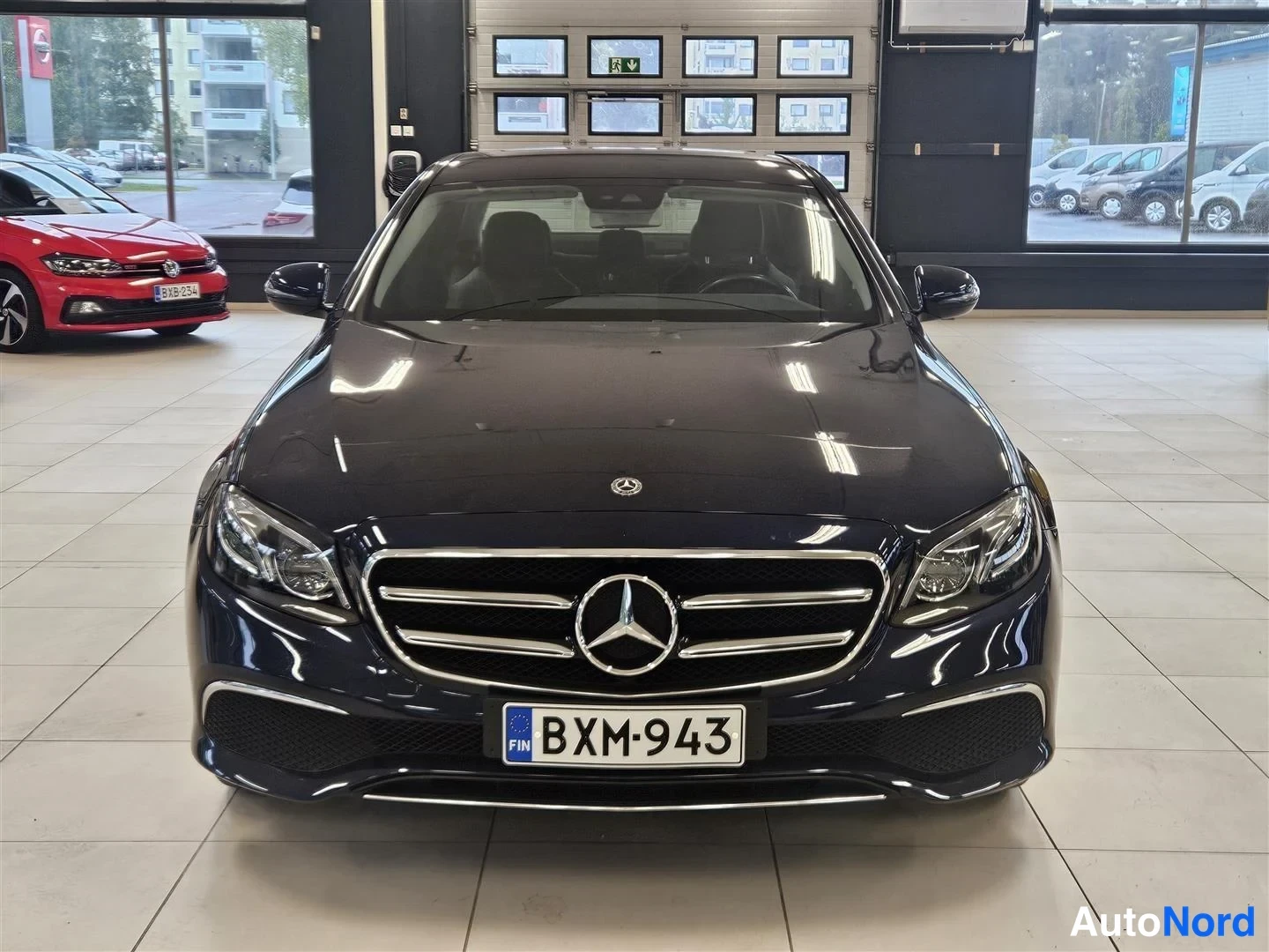 Edestä: Tummansininen Mercedes-Benz E-sarjan sedan suoraan edestä kuvattuna autoliikkeen tiloissa, rekisteritunnus.