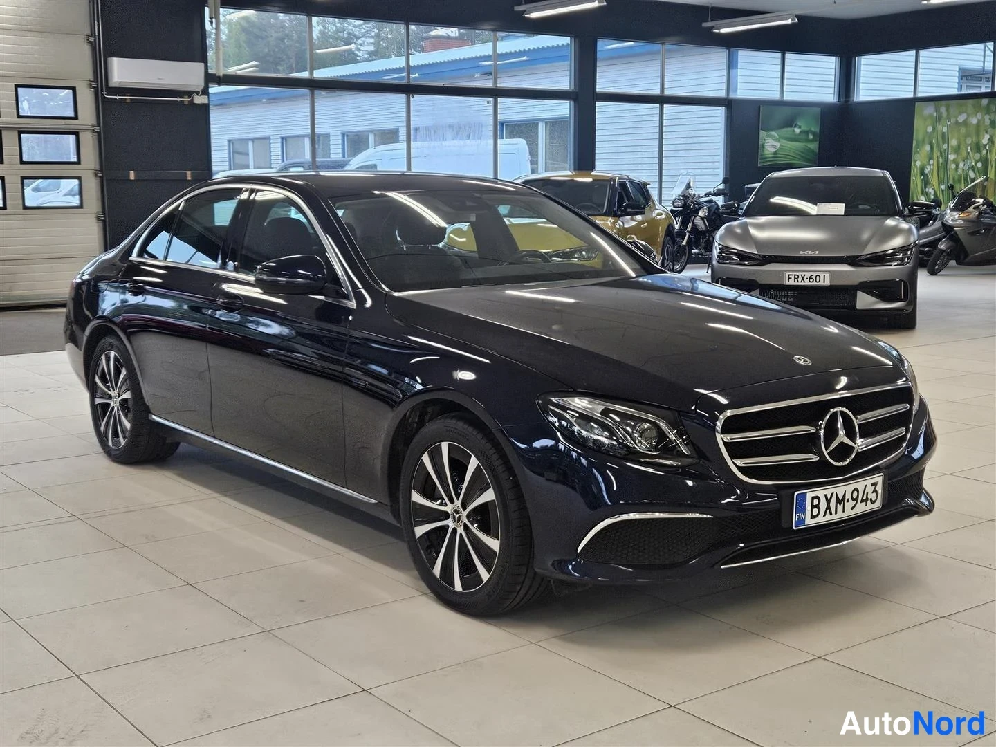 Etuviisto, nokka oikealle: Tummansininen Mercedes-Benz E-sarjan sedan edessä ja oikealta kuvattuna autoliikkeen showroomissa, rekisteritunnus.