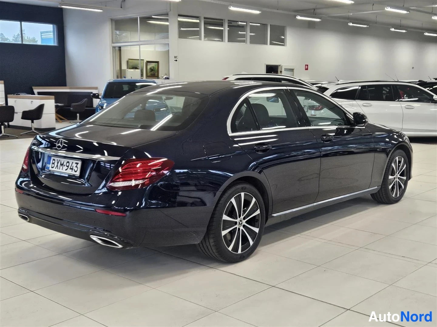 Takaviisto: Tummansininen Mercedes-Benz E-sarjan sedan takaa ja oikealta kuvattuna autoliikkeen showroomissa, rekisteritunnus.