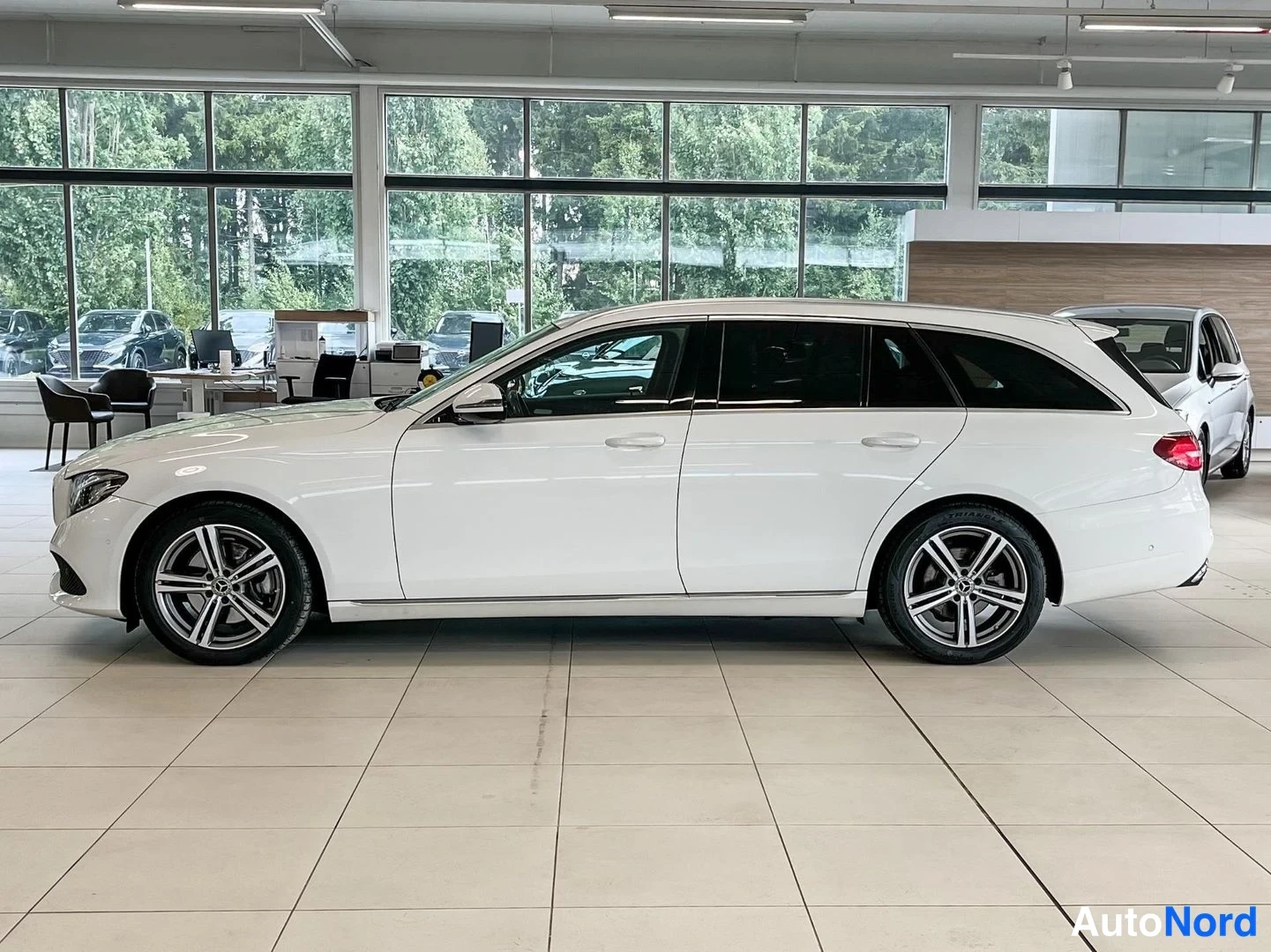 Sivulta: Valkoisen Mercedes-Benz E-sarja farmarin sivuprofiili autonhallissa, kylki ja molemmat pyörät näkyvät, takavalo osin näkyvissä.
