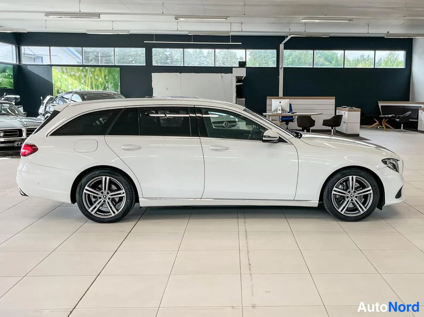 Sivulta: Valkoinen Mercedes-Benz E-sarja farmari sivuprofiilista autonhallissa, takapää ja kylki esillä, takavalo punainen.