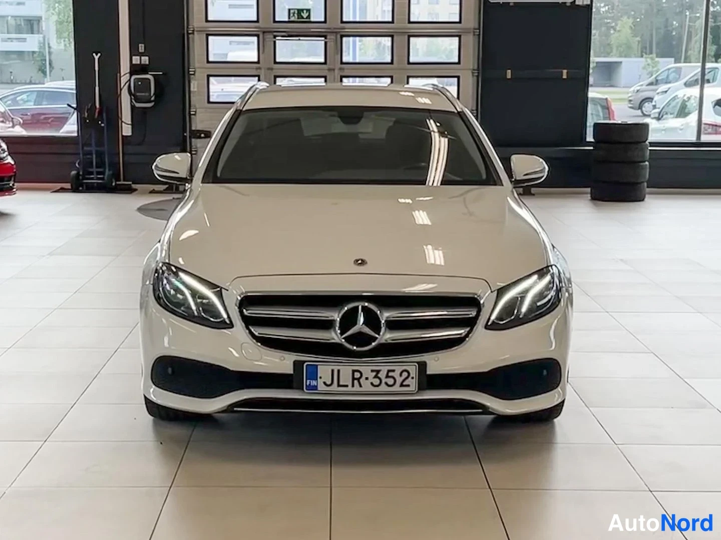 Edestä: Valkoinen Mercedes-Benz E-sarja suoraan edestä nähtynä autonhallissa, etumo hilarit ja tähtimerkki keskeällä, rekisterikilpi.