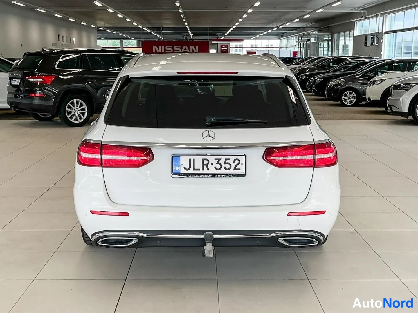 Takaa: Mercedes-Benz farmarin takapää suoraan takaa valkoisena autosalissa, rekisterikilpi, vetokoukku ja kaksoispakoputket näkyvissä.