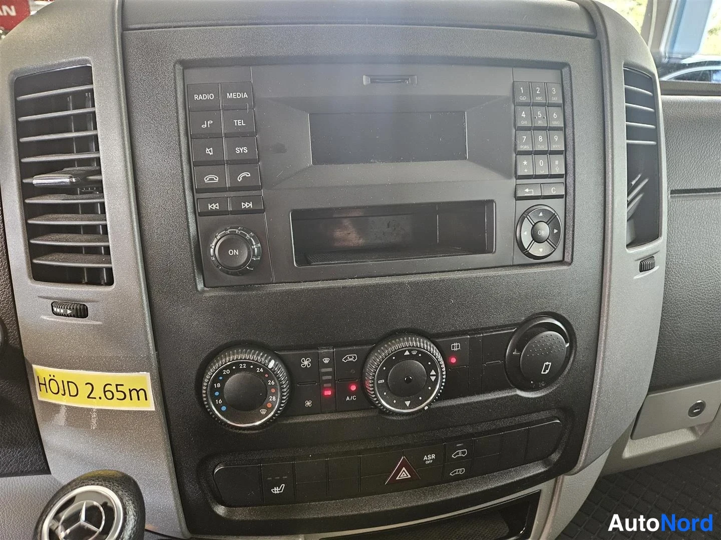 Muu: Keskikonsolin lähikuva: radio- ja ilmastointipaneeli A/C-merkinnällä, vieressä keltainen tarra 'HÖJD 2.65m'.