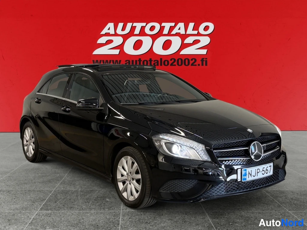 Etuviisto, nokka oikealle: Musta Mercedes-Benz A -henkilöauto etuviistosta oikealta, rekisteritunnus, panoraamakatto, punainen taustaseinä.