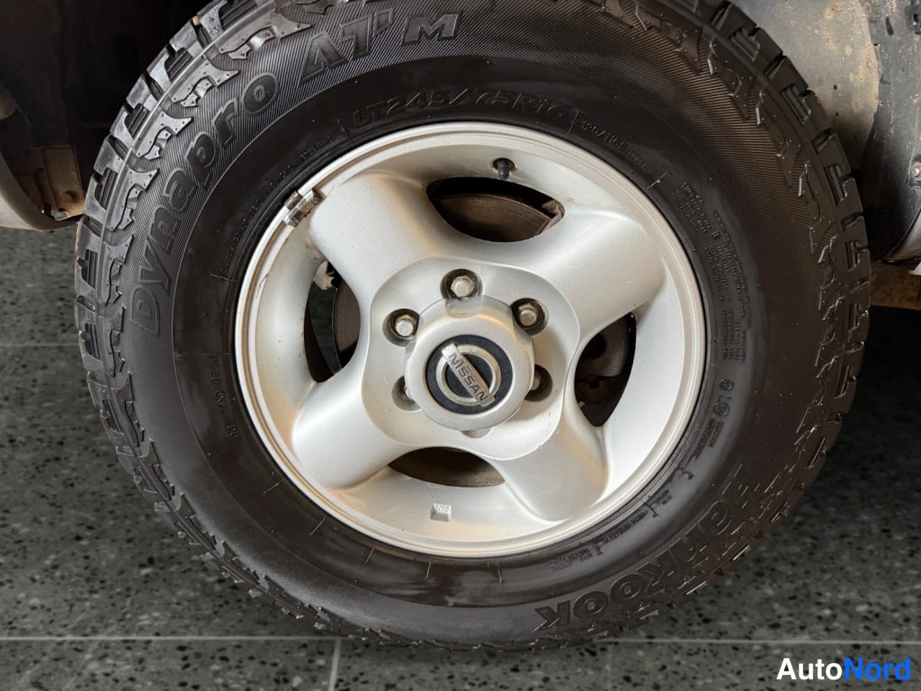 Yksityiskohta: Lähikuva auton pyörästä: hopeinen viisipulttinen Nissan-vanteen keskiosa, musta Hankook Dynapro ATm -renkas, jonka sivussa lukee LT245/75R16.