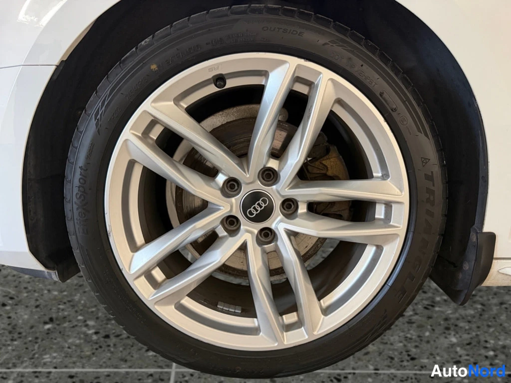 Yksityiskohta: Auton valkoinen alumiinivanne ja kesärenas lähikuvassa, Audi-merkki napassa, jarrulevy ja mäntä näkyvissä.