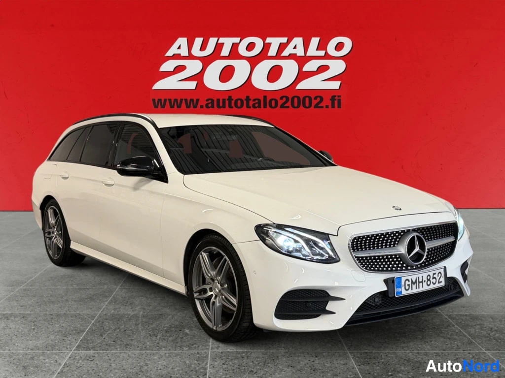 Etuviisto, nokka oikealle: Valkoinen Mercedes-Benz farmariauto kylkiview- ja etunakokulmassa punaisella taustalla.