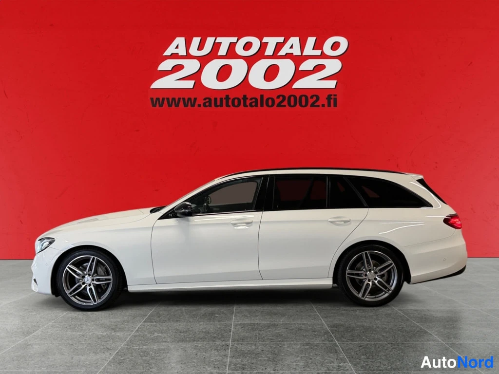 Sivulta: Mercedes-Benz E -farmari sivulta, valkoinen koripaneeli ja mustat kevytmetallivanteet punaisen taustan edessä.