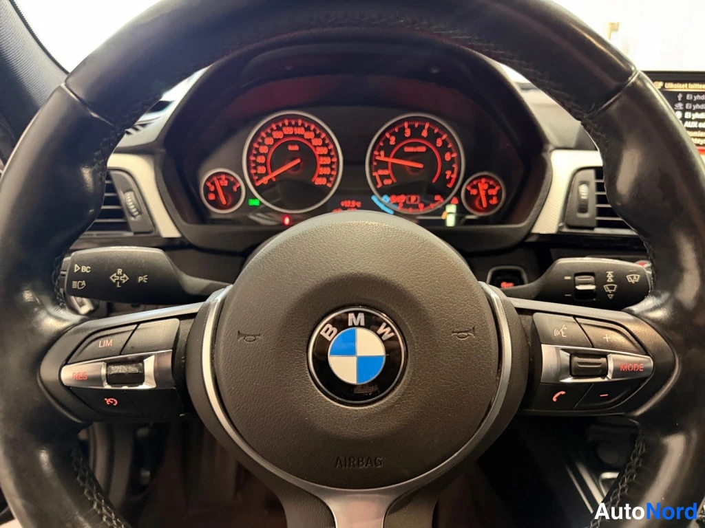 Muu: BMW-ohjauspyörä ilmatyynytäkstillä AIRBAG ja merkillä, taustalla punaisin valaistut analogiset mittarit ja ohjainnapit kuten LIM, RES, BC, MODE.