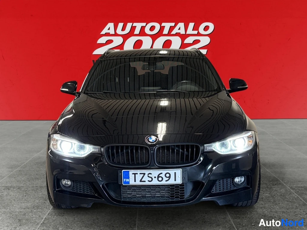 Edestä: Mustan auton BMW etupää suoraan edestä punaisella taustalla; ajovalot päällä ja rekisteritunnus.