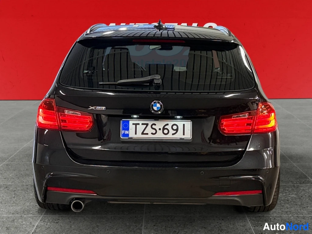 Takaa: Mustan auton BMW takapää suoraan takaa punaisella taustalla; rekisteritunnus ja pakoputki vasemmalla.