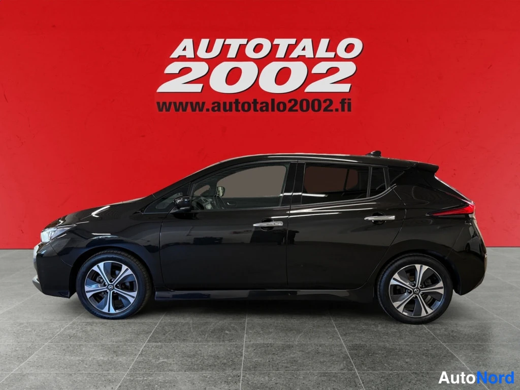 Sivulta: Nissan Leaf 2021, sivukuva, musta viistoperä seisoo harmaalla laatoitetulla lattialla punaista taustaa vasten.