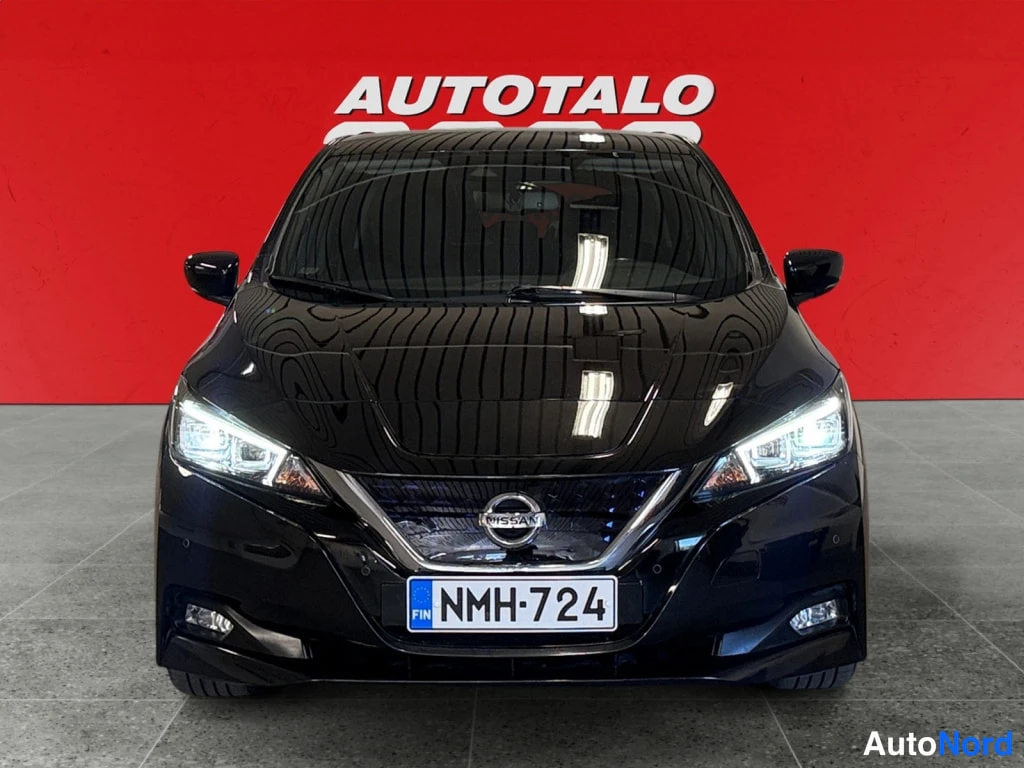 Edestä: Nissan Leaf 2021 suoraan edestä, otsavalot päällä, rekisteritunnus, musta kori punaisen taustan edessä.
