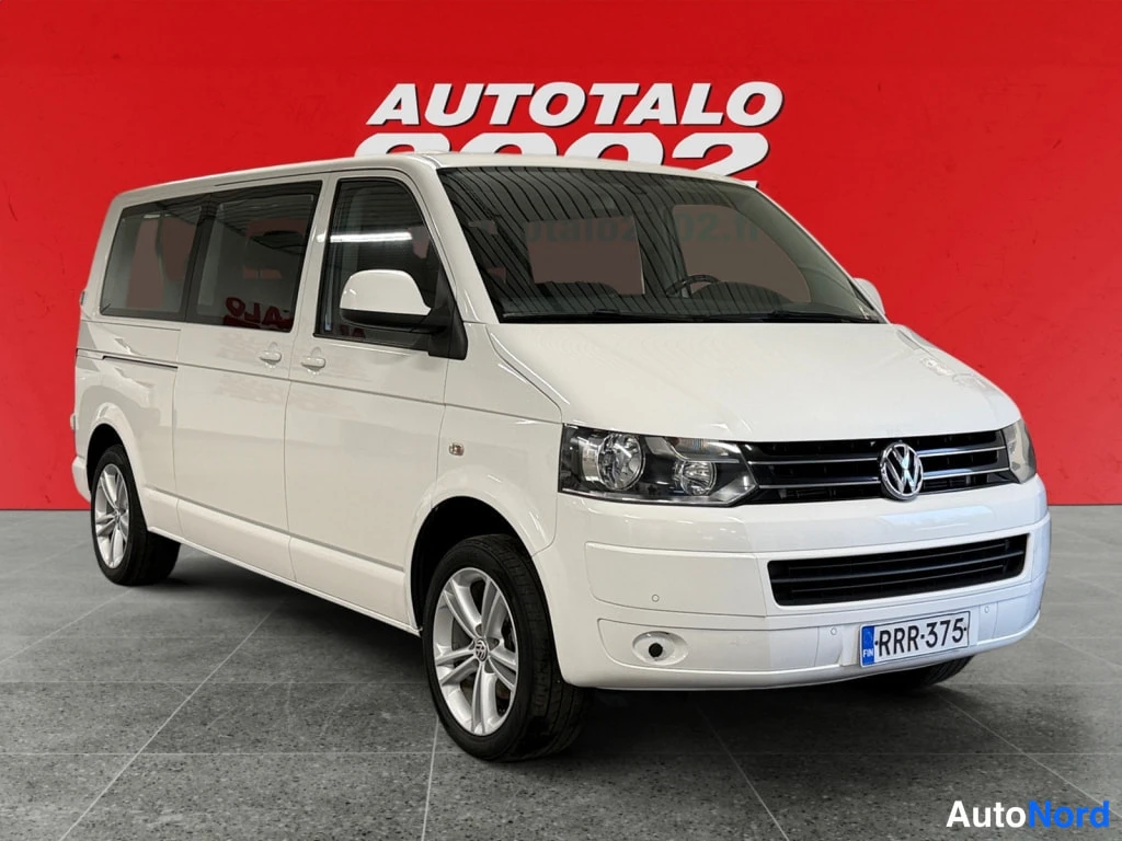 Etuviisto, nokka oikealle: Valkoinen Volkswagen Caravelle -pakettiauto etukulmasta, rekisteritunnus, punainen taustaseinä.