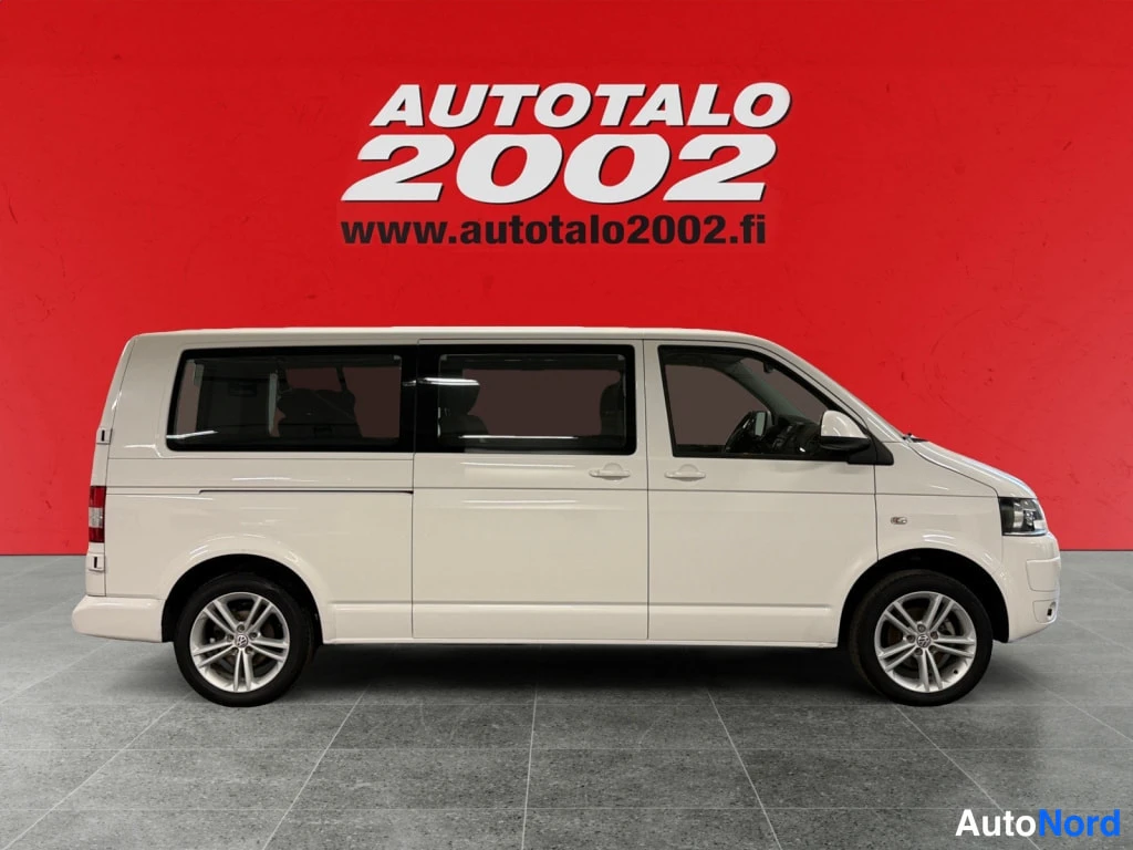 Sivulta: Valkoisen Volkswagen Caravelle -pakettiauton sivuprofiili, pitkä akseliväli ja kolme sivuikkunaa, punainen taustaseinä.