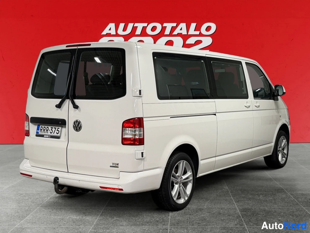 Takaviisto: Valkoisen Volkswagen Caravelle -pakettiauton taka- ja sivukulma, rekisteritunnus, TDI-merkintä ja veto-kytkin, punainen taustaseinä.