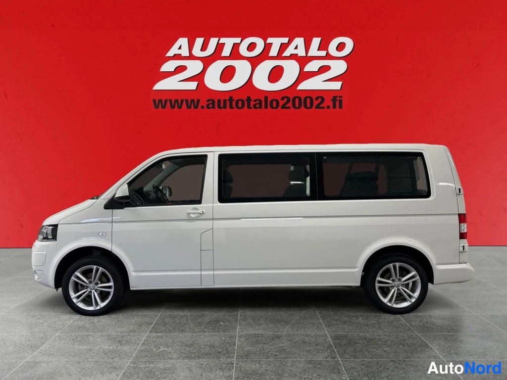 Etuviisto, nokka vasemmalle: Valkoinen Volkswagen Caravelle -pakettiauto sivulta etukulmasta, painettu kylki ja etupyörät näkyvät punaisen seinän edessä.