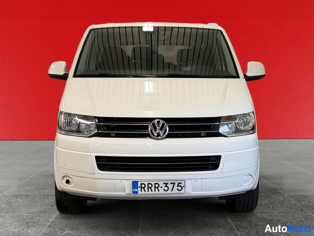 Edestä: Valkoisen Volkswagenin etunäkymä, ajovalot, konepelti ja rekisterikilpi punaisen taustan edessä.