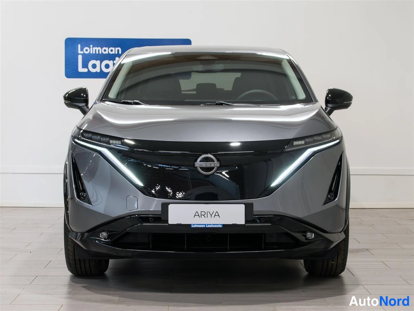 Edestä: Nissan Ariya, etuosa suoraan edestä; harmaassa korissa musta etusäleikkö, ohuet LED-ajovalot ja ARIYA-kyltti etupuskurissa.