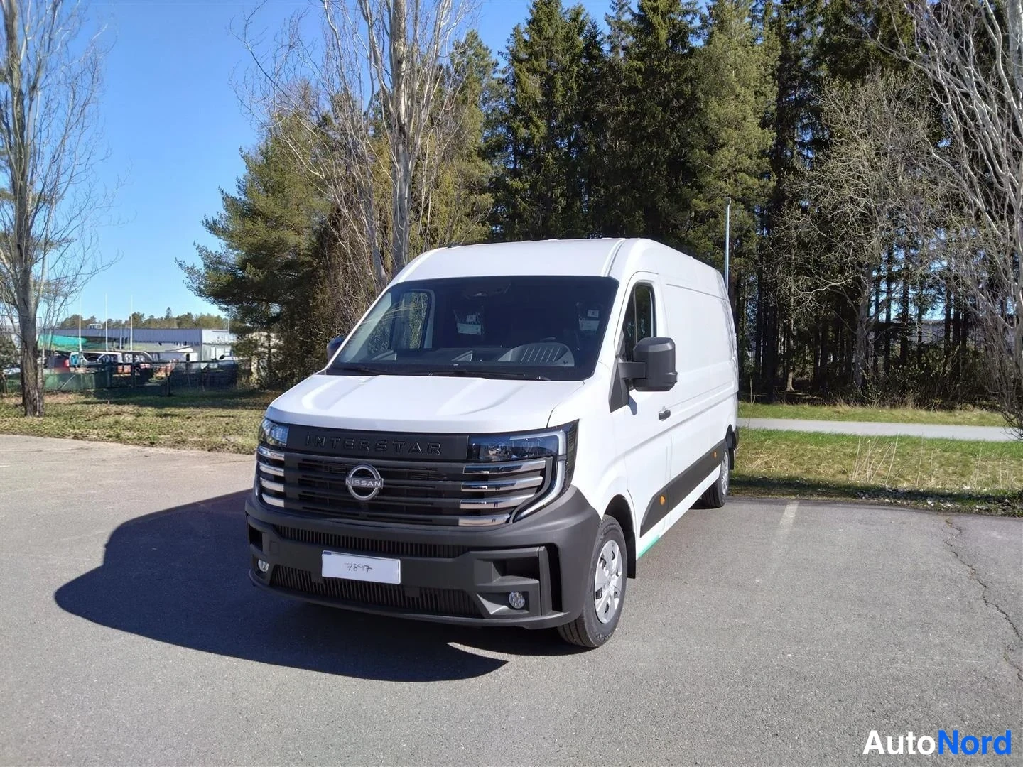 Etuviisto, nokka vasemmalle: Valkoinen Nissan Interstar -pakettiauto edestä vasemmalta kuvattuna parkkipaikalla puiston laidassa.