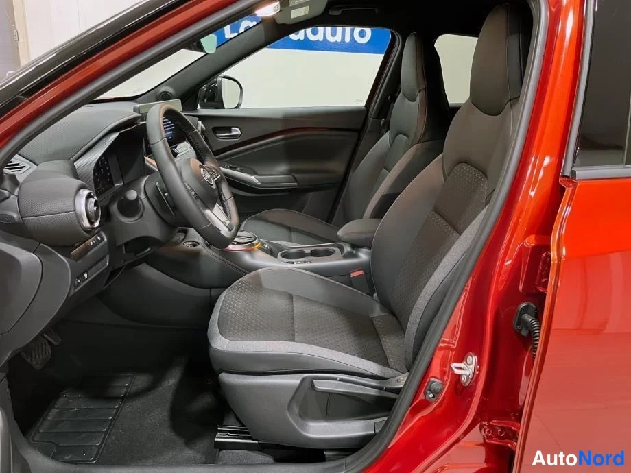 Sivulta: Nissan Juke:n etuistuin ja ohjaamo avoimesta vasemmasta etiovesta kuvattuna, tummat kankaatupit ja punainen korimaali näkyvät.