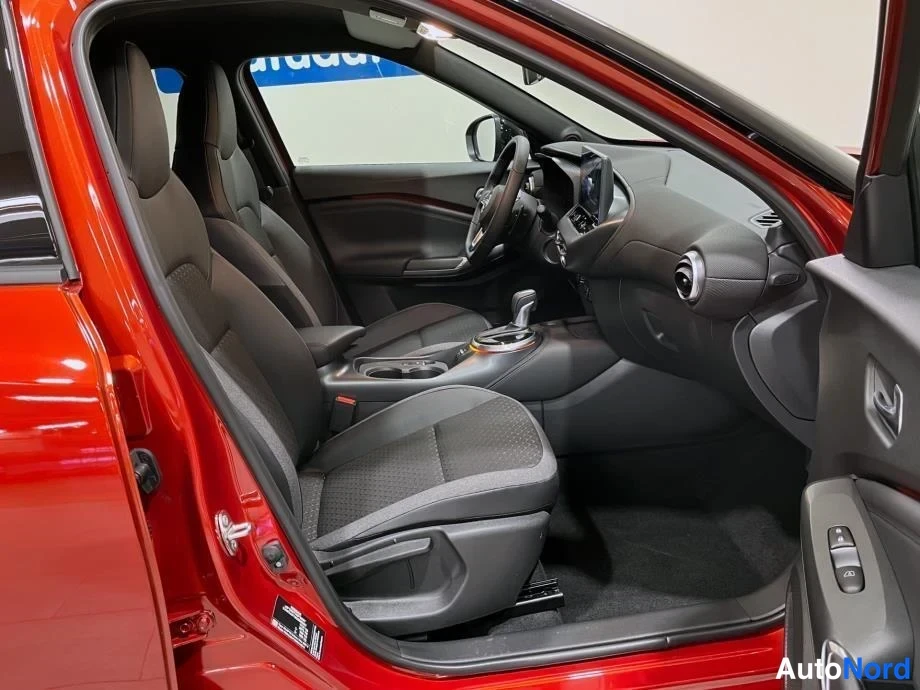Sivulta: Nissan Juke:n etuistuimet ja kojelauta avoimesta vasemmasta etiovesta kuvattuna, automaattivaihteiston valitsin ja kosketusnäyttö näkyvät.