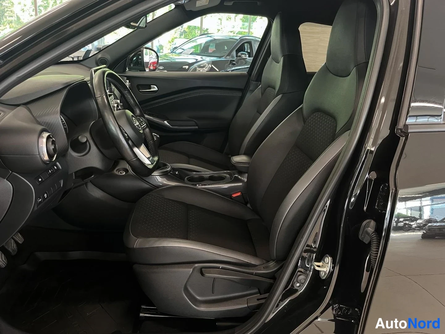 Muu: Nissan Juken ohjaamo kuljettajan puolelta: mustat urheiluistuimet, ohjauspyörä ja keskikonsoli, auto sisätiloissa näyttelyhallissa.