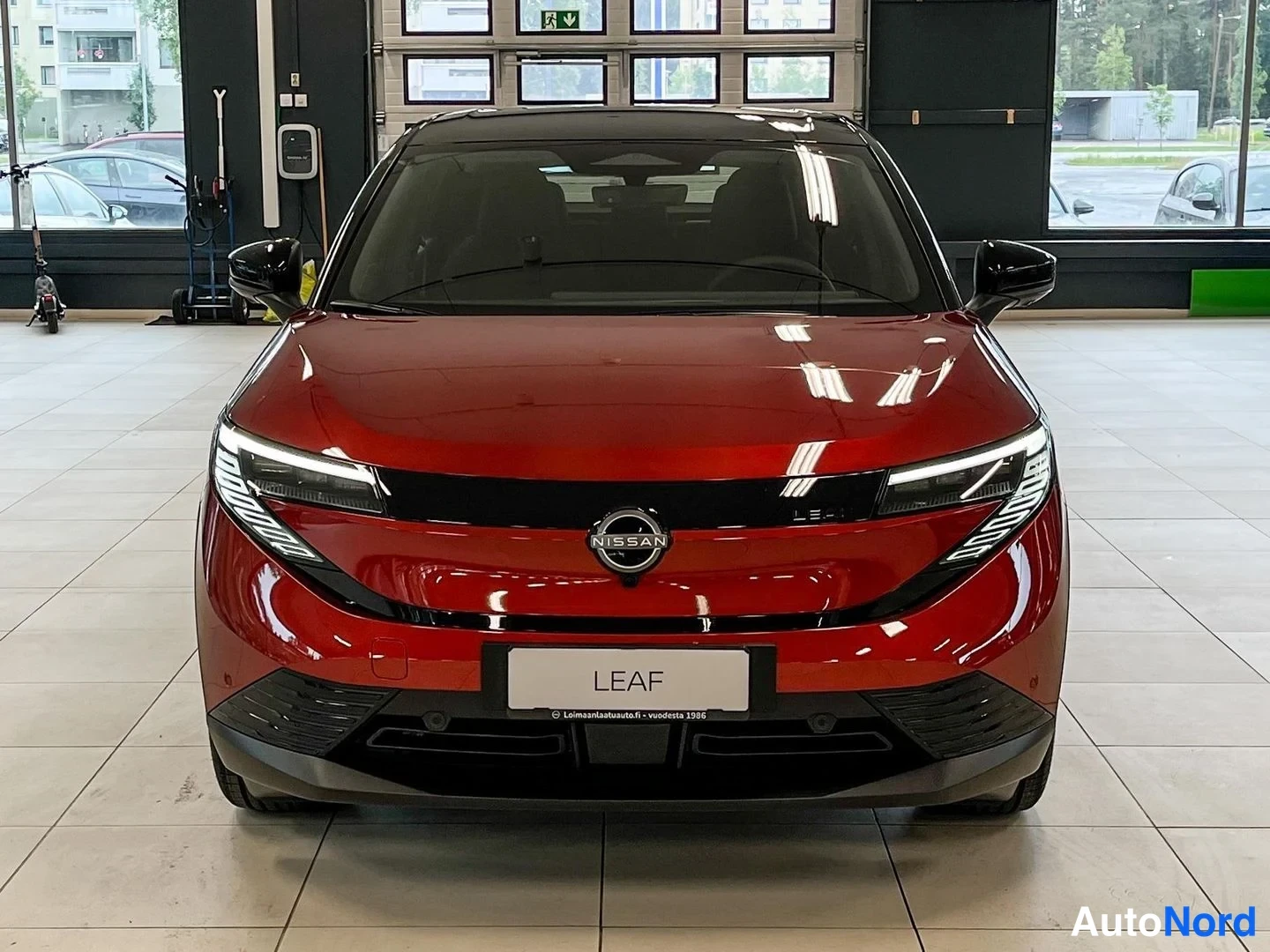 Edestä: Punainen Nissan Leaf suoraan edestä nähtynä autonhallissa, Nissan-merkki keulalla ja LEAF-teksti etukilvessä.