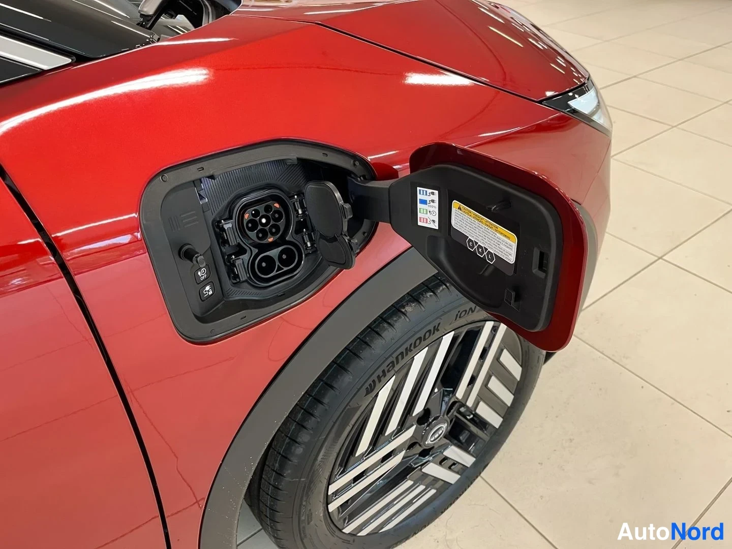 Sivulta: Punaisen auton avattu latausluukku etuosassa: sisällä Combined Charging -latauspistoke ja pienempi suuttimen kiinnike, luukun sisäpuolella varoituslappu, Hankoo