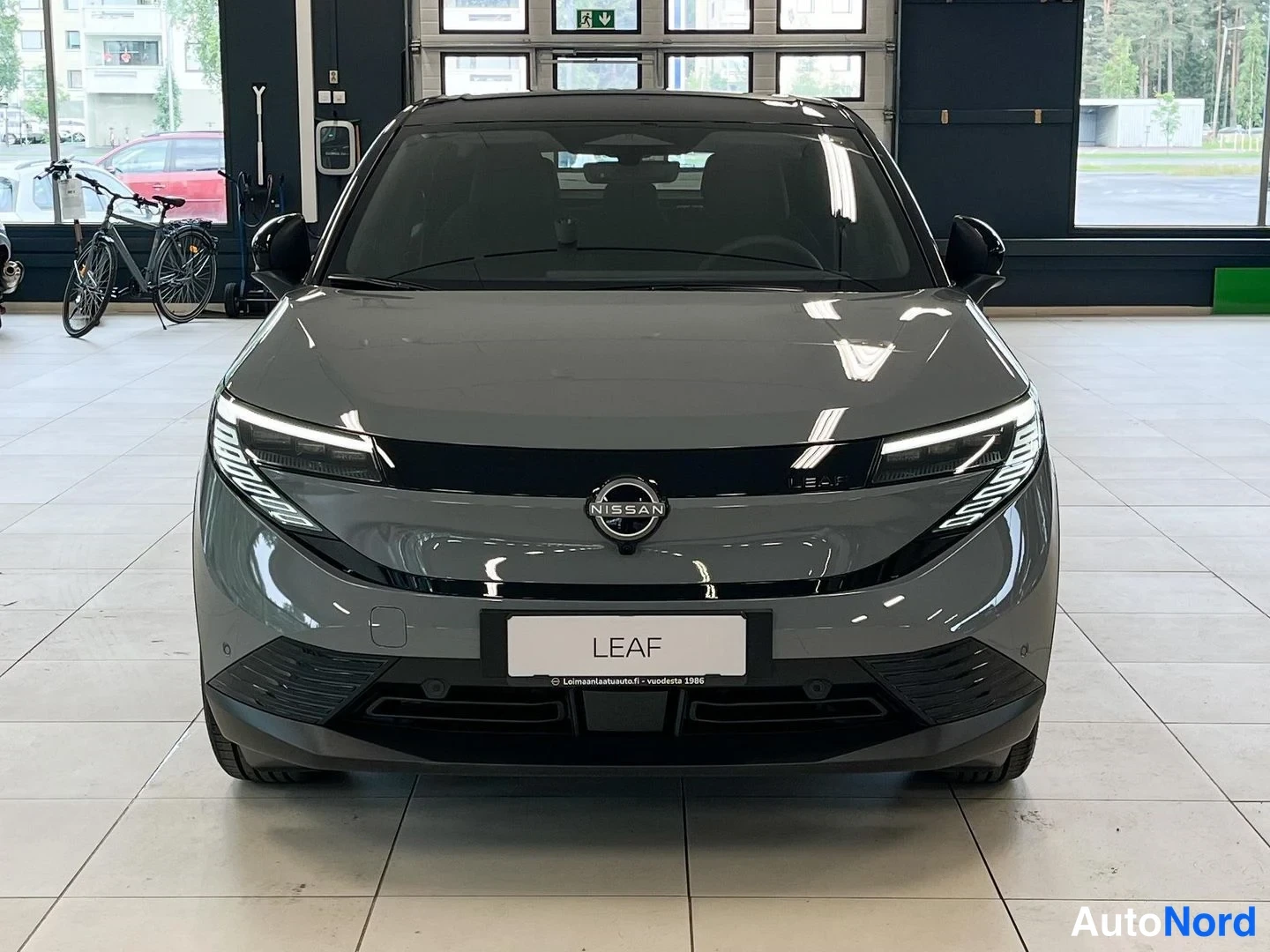 Edestä: Front view of a grey Nissan Leaf. The front grille features the Nissan logo and the license plate reads LEAF.
