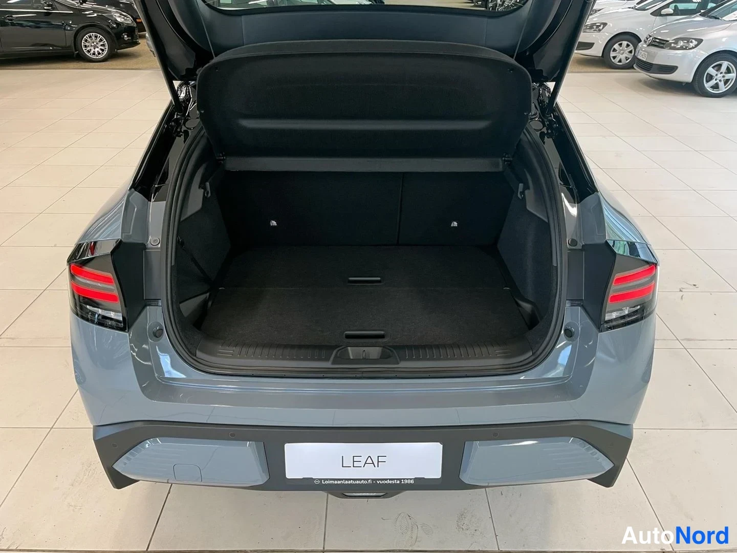 Tavaratila: Rear cargo area of a grey Nissan Leaf shown with an open trunk. A black parcel shelf is attached to the top of the opening.